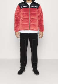 Červená sametová bunda s černou horní částí s nápisem "KARL KANI" v bílém textu, předním zipem a nadýchaným střihem, doplněná černými kalhotami a teniskami.
