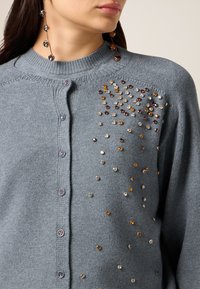 Lysegrå strikket cardigan med runde knapper og udsmykket med spredte perler i guld-, sølv- og bronzetoner på venstre side.