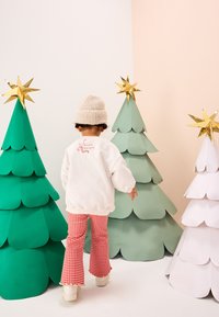 Felpa bianca con scritta rossa, pantaloni a zampa rossi a quadretti vichy, berretto crema. Circondato da alberi di Natale di carta verdi e bianchi con stelle dorate.