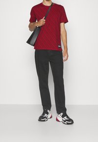 Camiseta roja con un patrón de bucles negros, combinada con pantalones negros y zapatillas negras y blancas. Bolsa de mano negra llevada al hombro.