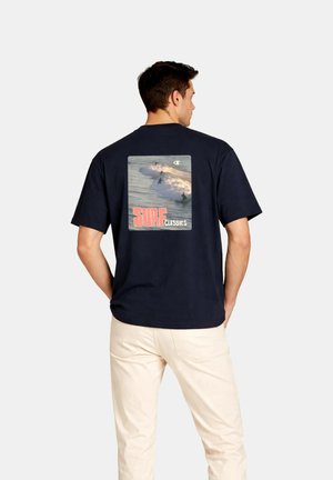 Mann trägt dunkelblaues T-Shirt mit Grafik von Surfern, die auf Wellen reiten, und dem Text „SURF CLASSICS“ auf dem Rücken, kombiniert mit beigen Hosen.