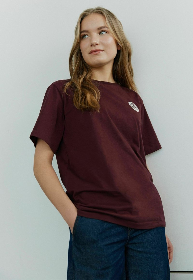 Bordeaux-rotes Baumwoll-T-Shirt mit kurzen Ärmeln und rundem Halsausschnitt, versehen mit einem kleinen runden Logo auf der Brust. Getragen mit dunklen Jeans.
