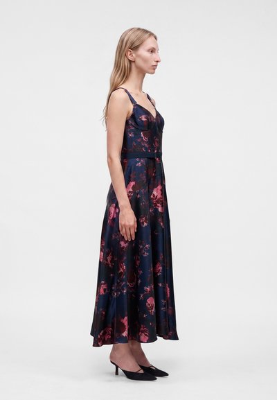 Femme debout de profil, portant une robe midi sans manches à fleurs foncées avec des fines bretelles et des chaussures noires à talons bas sur un fond uni.