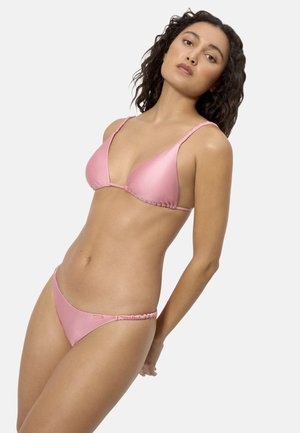 Mujer con cabello oscuro y ondulado, vestida con un bikini de dos piezas rosa, posando frente a un fondo liso de color gris claro.