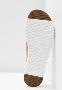 Semelle de chaussure UGG de couleur beige et blanche avec une adhérence texturée, talon et embouts marron, et logo UGG embossé sur la partie du talon.