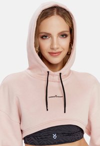 Roze skraćeni dukserica od mekog materijala s kapuljačom na vezanje i crnim logotipom "CARDIO BUNNY". Nosena preko tamnog sportskog grudnjaka.