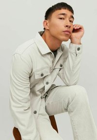 Giacca in denim grigio chiaro con chiusure a bottoni e tasche sul petto, abbinata a pantaloni grigio chiaro coordinati, caratterizzati da una texture liscia.