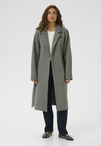 Pelēks vilnas mētelis ar griezuma apkakli, vienu pogu un garām piedurknēm. Valkāts virs baltas blūzes un kopā ar tumšiem džinsiem un rakstainiem apaviem.