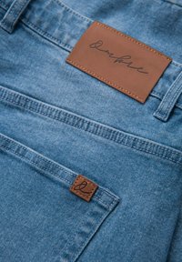 Blaue Jeans aus Denim mit Gesäßtasche, die ein kleines Lederpatch und ein größeres Lederetikett zeigt, das nahe am Bund angenäht ist.