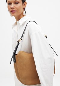 Borsa a spalla in suede tan con forma curvata, tracolla in pelle nera e dettagli in metallo dorato, posizionata su uno sfondo bianco.