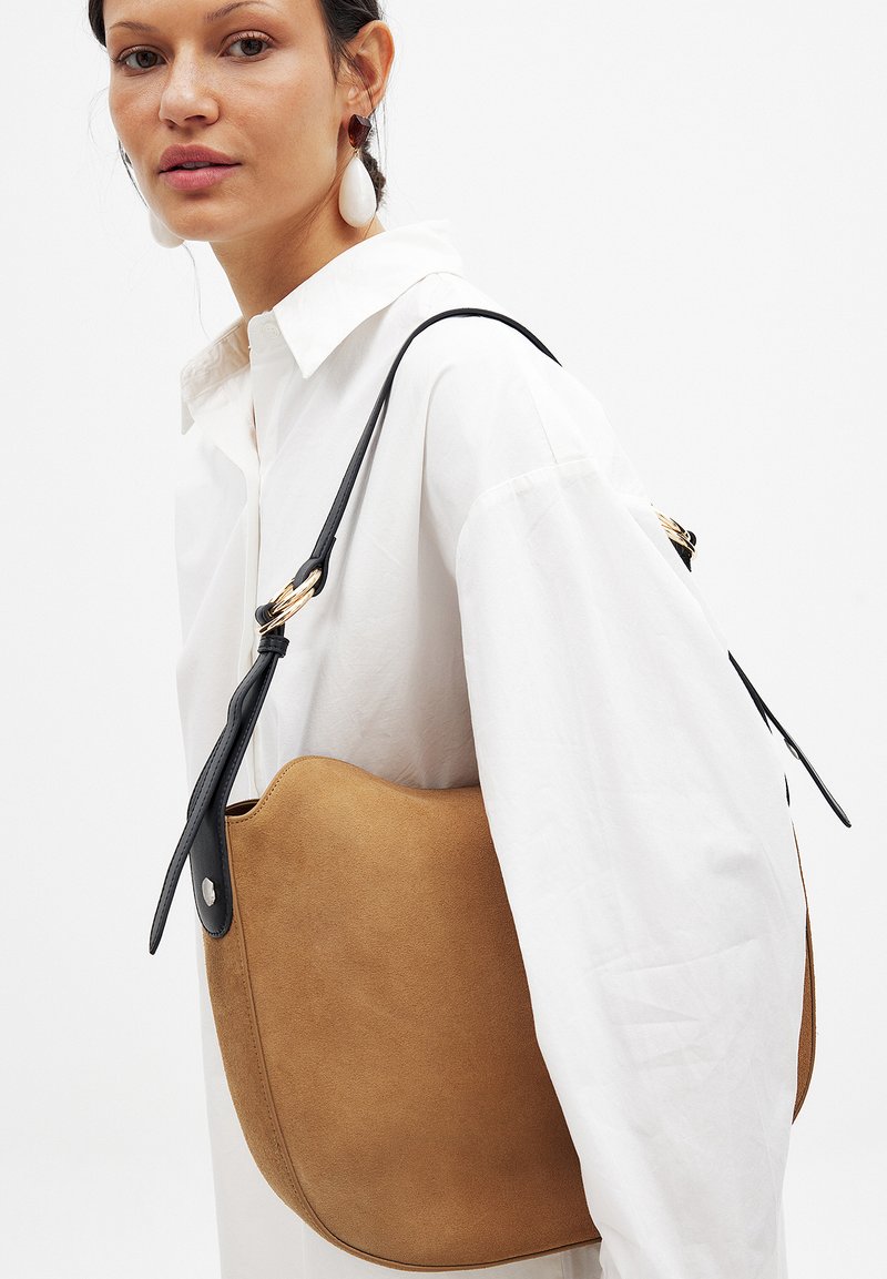 Borsa a spalla in suede tan con forma curvata, tracolla in pelle nera e dettagli in metallo dorato, posizionata su uno sfondo bianco.