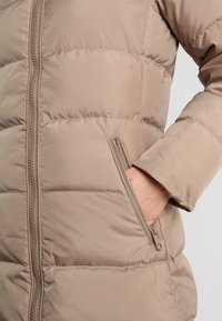 Hnědá péřová bunda s prošívaným designem, zapínáním na zip a boční kapsou. Textura vypadá měkce a izolovaně pro pohodlí a teplo.