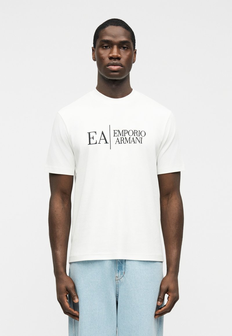 Άνδρας που φοράει λευκό κοντομάνικο μπλουζάκι Emporio Armani και ανοιχτό μπλε τζιν, στέκεται μπροστά σε απλό φόντο.