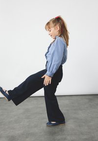 Blauwe en witte blouse met patroon en pofmouwen, gecombineerd met marineblauwe wijde broek en marineblauwe flats, staande op een grijze vloer.