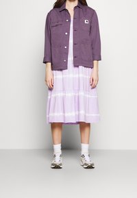 Lila bomullsjacka med knappstängning och två framfickor, lager på lager över en ljuslila tie-dye klänning med vågig fåll. Avslutad med sneakers.