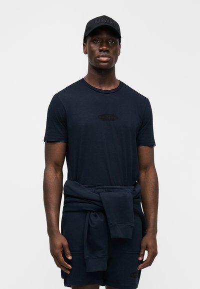 Černá čepice s logem "VOLTAIRE", tmavě modré tričko a šortky. Bavlněná látka, volný střih. Svetr uvázaný kolem pasu ve stejné barvě. Jednoduchý design.