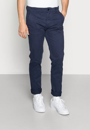 Hombre con chinos azul marino remangados y zapatillas blancas, de pie con una mano en el bolsillo contra un fondo claro y liso.