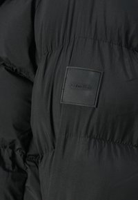 Čierna nafukovacia bunda s hladkou textúrou a prešívaným dizajnom. Obsahuje vyvýšenú čiernu obdĺžnikovú nálepku s logom "Calvin Klein".