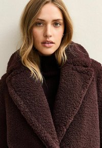Giovane donna con capelli biondi indossa un cappotto oversize strutturato marrone scuro sopra un dolcevita nero, guardando direttamente avanti.
