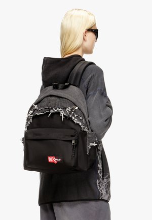 Mochila negra con acentos grises deshilachados en la parte superior, que cuenta con un bolsillo frontal y un parche con el logotipo. Fabricada con materiales duraderos, diseño estructurado.