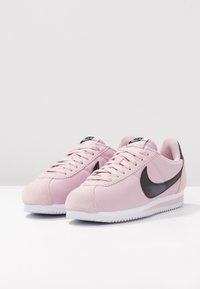 Nike-sneakers med ett pastellrosa textilöverdrag, svart swoosh-logotyp och vit gummisula med zigzag-mönster.