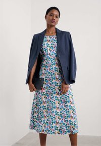 Robe fleurie avec des motifs violets, bleus et jaunes, associée à un blazer navy. Mannequin tenant une pochette noire. Tissu lisse et design à hauteur du genou.