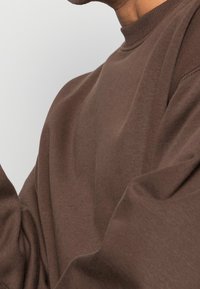 Felpa marrone con scollo rotondo e maniche lunghe, realizzata in tessuto morbido e strutturato. Dettagli di cuciture visibili sulle maniche.