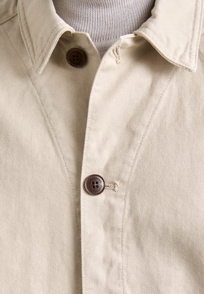 Chaqueta beige de botonadura con un cuello clásico, detalles de botones marrones y una tela texturizada y duradera. Sin patrones ni acentos visibles.