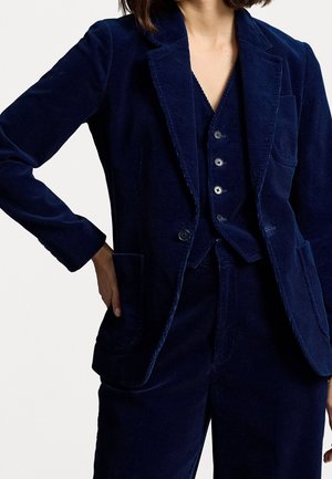 Frau trägt einen dunkelblauen Cord-Blazer, passende Weste mit Knöpfen und Hose, mit einer Hand in der Tasche vor schlichtem Hintergrund.