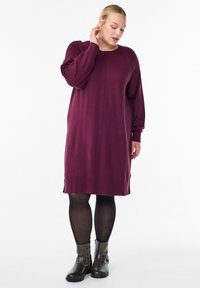 Žena u haljini dužine do koljena s dugim rukavima u šljivinoj boji, crnim tajicama i tamnim gležnjačama, stoji i dodiruje vrat.