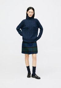 Pulover din lână de culoare bleumarin, cu guler înalt, asortat cu o fustă mini în carouri de culoare verde închis și albastru, completat de șosete închise la culoare și pantofi tip loafers.