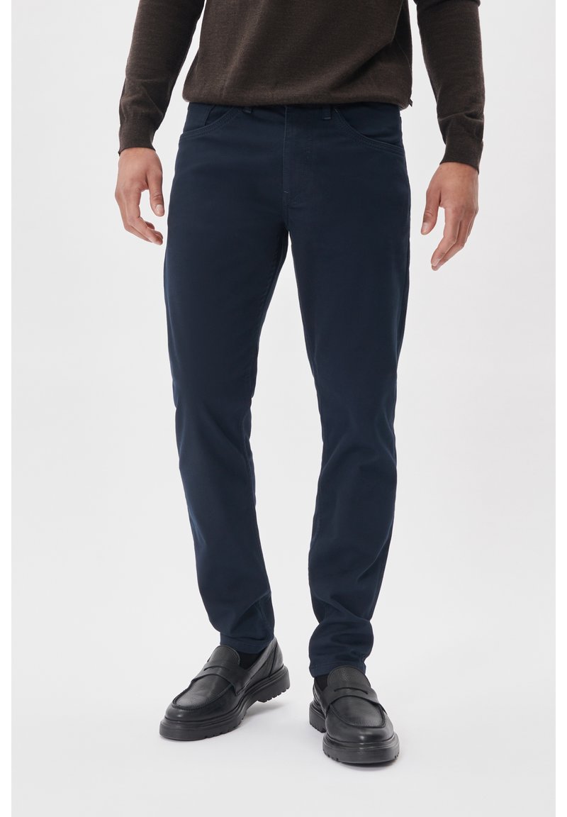 Homme portant un pantalon slim bleu marine foncé, un pull marron foncé et des mocassins en cuir noir, debout sur un fond blanc.