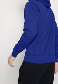 Felpa blu con maniche lunghe, realizzata in un materiale morbido. Presenta una tasca a marsupio e polsini a coste. Indossata con pantaloni neri.