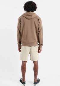 Sweat à capuche marron de coupe décontractée, assorti à un short beige clair avec une poche arrière. Le modèle est vu de dos, portant des chaussures noires sans lacets.