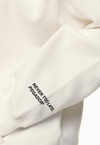 Manica di una felpa bianca con testo nero "NEVER TOO LATE. PEGADOR" vicino al polsino, che mostra la texture del tessuto e i dettagli del polsino.