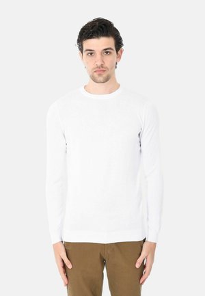 Giovane uomo con capelli scuri che indossa una maglietta bianca semplice a maniche lunghe e pantaloni marroni, in piedi davanti a uno sfondo grigio chiaro.