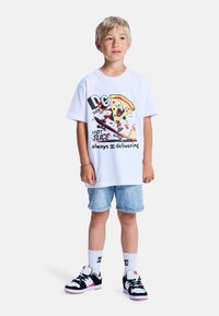 Ein weißes Baumwoll-T-Shirt mit einem bunten Grafikdesign eines Cartoon-Pizzastücks auf einem Skateboard. Kombiniert mit Jeansshorts und schwarzen Sneakers.