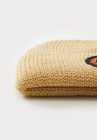 Bonnet en tricot de couleur jaune clair, avec un motif texturé et une patch ronde noire avec des accents colorés sur le côté.