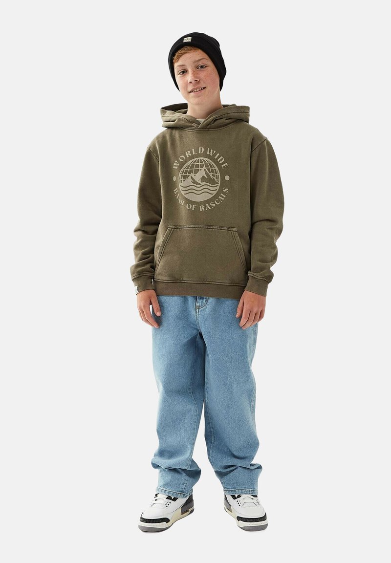 Olivgrön hoodie med grafiskt tryck, känguruficka, i kombination med lösa blå jeans och svartvita sneakers. Svart mössa på huvudet.