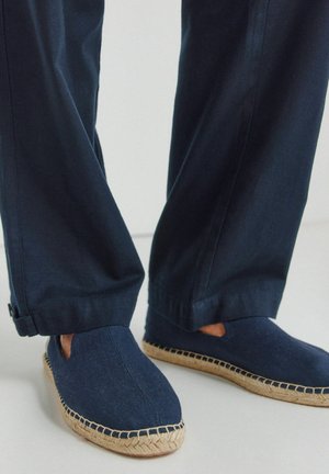 Persoon draagt donkerblauwe espadrille instapschoenen met gevlochten jute zolen en donkerblauwe broek, staand op een lichte vloer.