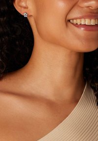 Blauwe bloembloemvormige oorbellen met een centrale edelsteen, geplaatst in een kleine metalen basis, gedragen aan het oor van een model, die een geribbelde top aanvult.