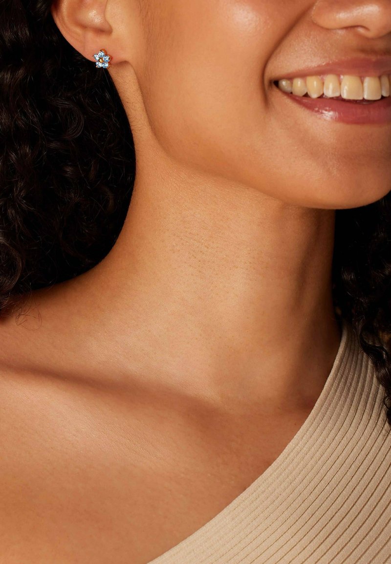 Blauwe bloembloemvormige oorbellen met een centrale edelsteen, geplaatst in een kleine metalen basis, gedragen aan het oor van een model, die een geribbelde top aanvult.