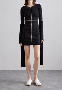 Svart ribbad klänning med långa ärmar och guldstickning fram; matchad med vita träskor. Klänningen har en figurnära passform.