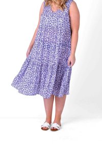 Woman wearing a sleeveless, blue floral midi dress with white sandals, standing against a white background.