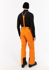 Persoon in een zwart langetermijnshirt, zwarte beanie, oranje skibroek met bretels en zwarte skischoenen, staande met de rug naar de camera op een witte achtergrond.