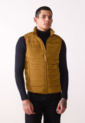 Puffy, mustard-yellow vest with a high collar and zip closure. The fabric is smooth with horizontal stitching; layered over a black turtleneck.