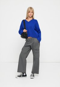 Plavi oversized pulover, sivi cargo hlače s bočnim džepovima i crne Converse tenisice. Model nosi malu crnu torbicu preko ramena.