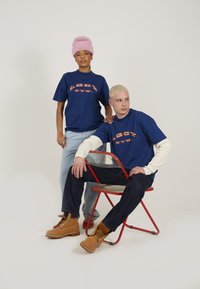 Blå grafiske t-shirts med orange tekst, afslappede jeans og lyse støvler. En model bærer en pink fluffy hue og sidder på en rød stol.