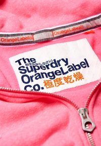 Sudadera rosa con cremallera frontal, etiqueta de tela con texto en azul y naranja, y una etiqueta interior marcada "OrangeLabel."