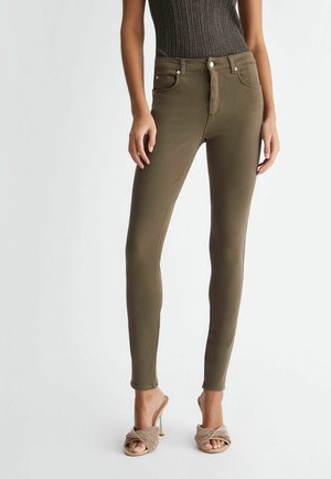 Dopasowane oliwkowozielone dżinsy noszone z beżowymi, wysokimi sandałami z plecionki i ciemnym, bezrękawnikowym topem na białym tle.